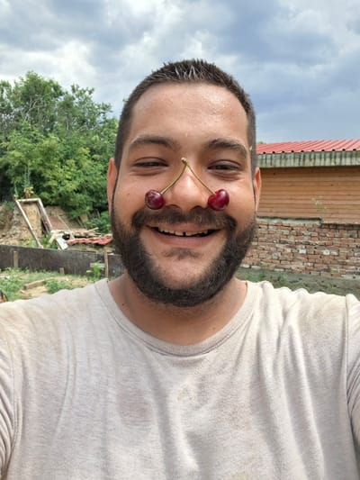 The media file is a selfie taken outdoors in a rural setting. The primary subject is a man with dark hair and a beard, who is smiling and looking at the camera. He is wearing a light-colored t-shirt. Two cherries are placed on his nose, with their stems visible.
The background of the image shows a brick wall and a wooden structure, likely part of a building. There are also trees and a cloudy sky, suggesting it is daytime. The overall atmosphere is casual and playful, indicated by the man's expression and the use of cherries as a prop.
The provided location context of Dolna Malina, Bulgaria, suggests the setting is a village or rural area in Bulgaria. There is no visible text in the image. The image is oriented vertically, with the man's head at the bottom and the sky at the top.