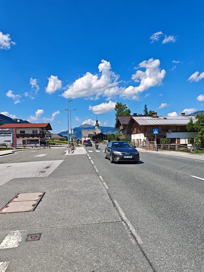 The image depicts a sunny day in Filzen, Austria, with a bright blue sky dotted with white clouds. A paved road runs through the scene, with a dark gray sedan driving on the right lane and a motorcycle on the left. On the left side of the road, there are buildings, including one with a red roof and a sign that reads "Ski ruft! GÄTTBAHN Comfortline LEITNER". Further down the road, a church with a steeple is visible, surrounded by traditional Austrian architecture. The landscape is mountainous, with green hills and trees in the background. The road has white lane markings, and there are streetlights and a pedestrian crossing sign. The ground on the left side of the road has manhole covers.