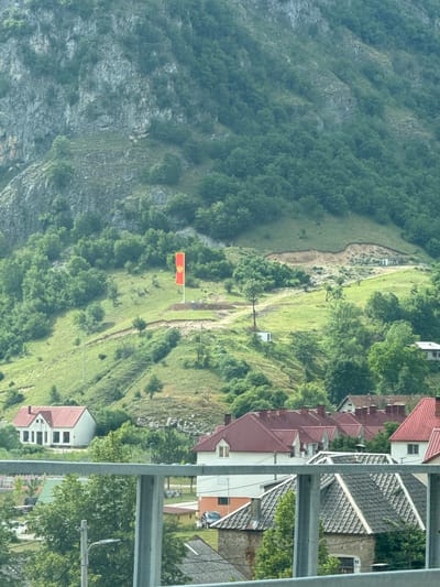 In Šavnik, Montenegro, a red and gold flag, likely the flag of Montenegro, is prominently displayed on a tall flagpole atop a lush green hillside. The flag stands against a backdrop of a steep, rocky mountain face covered in dense green forest. The hillside itself is a mixture of grassy slopes and trees, with a dirt path winding its way upwards. Below the flag, several small, white structures are visible, possibly utility sheds or outbuildings. Further down the slope, a cluster of houses with distinctive red roofs can be seen, nestled amongst the trees. In the foreground, a metal railing, part of a bridge or viewing platform, runs horizontally across the bottom of the image, partially obscuring a view of more houses with red and gray roofs and some vehicles. The overall scene depicts a rural mountainous landscape with signs of human settlement.