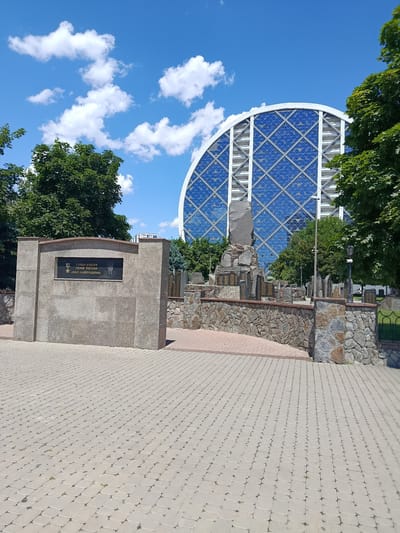 The image shows a memorial with a large granite monument on the left, inscribed with text and a portrait. To the right of this monument is a complex stone structure with numerous upright stone slabs, possibly commemorating individuals. In the background, a distinctive modern building with a semicircular, blue, diamond-patterned glass facade is visible. Lush green trees frame the scene, and the ground is paved with interlocking light-colored stones. The sky above is blue with scattered white clouds. This memorial is located in Grozny, Russia, specifically in Akhmat-Khadzhi Kadyrov Square, in front of the State Theatre of Nartai.