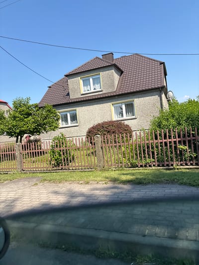 The image shows a two-story house with a brown tiled roof, situated in Bralin, Poland. The house has gray, textured walls and several windows, some with yellow trim. A red metal fence runs along the front of the property, separating it from a paved walkway and a grassy area. A small tree and bushes are visible in the yard. The sky is clear blue, indicating a sunny day. The image is taken from a slightly elevated perspective, possibly from a vehicle, as evidenced by the shadow and reflection in the foreground. There are no people or animals visible in the scene.