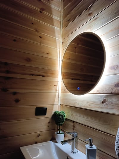 This is a photograph of a modern bathroom with wooden paneled walls. The primary subject is a large, oval mirror with integrated LED lighting, positioned in the corner of the room. The mirror reflects the warm tones of the wood and the surrounding light. Below the mirror, a white sink is visible, featuring a sleek chrome faucet. To the right of the faucet is a white soap dispenser. A small, potted artificial plant with a topiary-like shape sits to the left of the faucet. A black electrical outlet is mounted on the wooden wall above and to the left of the plant. The lighting suggests it is nighttime or the room is dimly lit, with the LED light around the mirror being the main source of illumination. The overall aesthetic is contemporary and clean, with a focus on natural materials and modern fixtures. There is no visible text in the image.