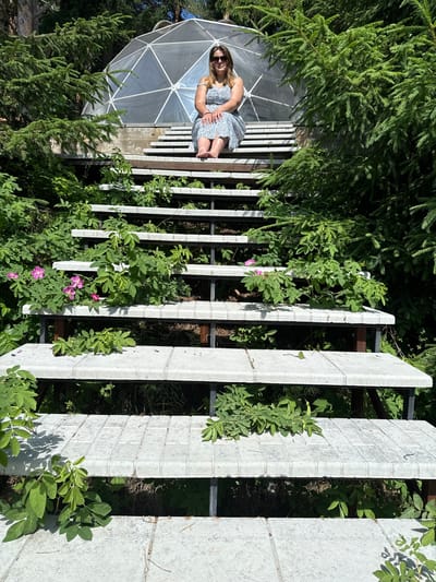 A woman with blonde hair and sunglasses is sitting on a set of concrete stairs. She is wearing a blue and white patterned dress. Behind her, a geodesic dome structure is visible. The stairs are surrounded by lush green foliage and trees. The scene appears to be outdoors. There is no information available to determine the location in Makarovo, Russia.