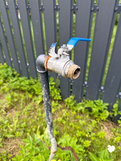An outdoor, close-up, eye-level shot depicts a water faucet attached to a black pipe. The faucet is made of silver-toned metal and has a blue lever handle. A white sticker with blue lettering is affixed to the faucet. The pipe is vertical and extends downwards out of frame. The background consists of a dark grey, slatted fence and lush green foliage. The scene is located in Tēraudi, Latvia.