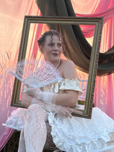 A young Caucasian woman with curly auburn hair and fair skin is pictured holding a white lace fan and a gold-colored frame. She is wearing a yellow, off-the-shoulder dress with ruffles, sheer white opera gloves, and white lace stockings. Her hair is styled up with small pearls. She is seated on a white bench with ruffles, and the background consists of draped pink and dark green fabric. The lighting suggests it is either dawn or dusk. There is no identifiable indication of the location within the city of Foki, Russia.