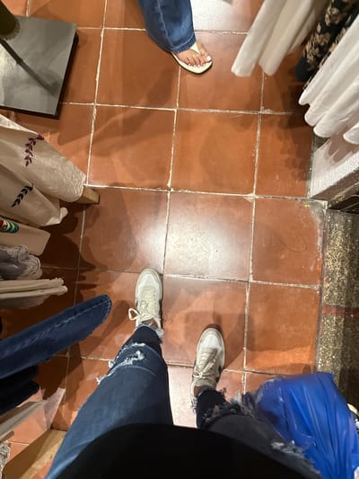The image shows an indoor scene of a retail space with terracotta tile flooring. The composition is a high-angle shot capturing the lower portions of three individuals and various objects on the floor. One person is wearing blue jeans and a sandal with white toenail polish. A second individual is wearing distressed denim jeans and white sneakers. A blue plastic bag sits near this person. The third person is wearing dark-colored jeans and white sneakers. Rolls of fabric are scattered on the floor, some appearing to be on stands. The scene's location is inferred to be a retail shop or market in San Cristóbal, Mexico, although there are no elements of the image to directly confirm this.
