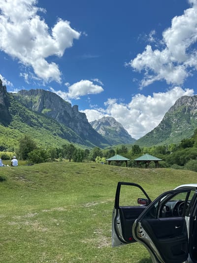 The image depicts a scenic mountainous landscape in Gusinje, Montenegro. In the foreground, a silver car with its doors open is parked on a grassy field. Two people, wearing white shirts and hats, are seated on the grass in the middle ground, facing away from the camera. Behind them, two green gazebos with pointed roofs are visible. The background is dominated by lush green mountains under a bright blue sky with scattered white clouds. The terrain is uneven with rolling hills and dense vegetation covering the mountain slopes.