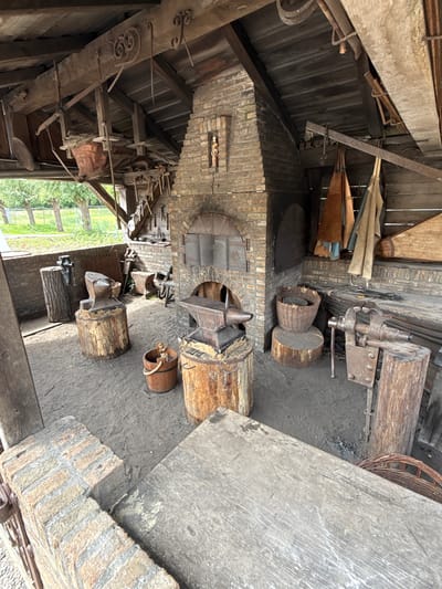 The image depicts the interior of a blacksmith shop. In the center is a brick forge with a chimney. Several anvils are mounted on wooden stumps. Various metalworking tools are present, including a vise and tongs. The structure is a wooden shed with a dirt floor. The scene is located in Eindhoven, Netherlands. There are no people visible in the image.