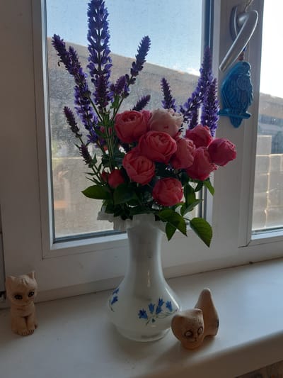 A bouquet of pink roses and purple lavender is displayed in a white vase on a windowsill. The vase has a blue floral pattern. Two small, light brown ceramic figures, one shaped like a cat and another like a bird, are placed near the vase. A silver window handle is visible in the upper left corner, along with a blue ceramic bird decoration. The scene appears to be indoors, with natural light coming through the window. The weather outside is unclear, but the light suggests it is daytime. The setting is a windowsill, likely in a home or apartment in Progress, Kyrgyzstan. No visible text is present.