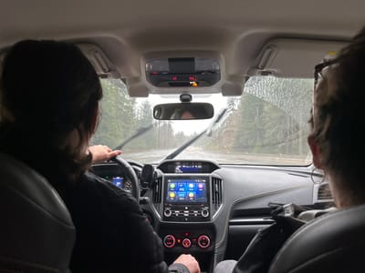 From inside a car, a driver is seen steering while windshield wipers clear the rain. A passenger sits beside the driver. Outside, trees line a wet road under an overcast sky. The dashboard is visible with a touchscreen display and climate controls. An interior mirror and overhead console are also in view. The road ahead appears slick and potentially hazardous.