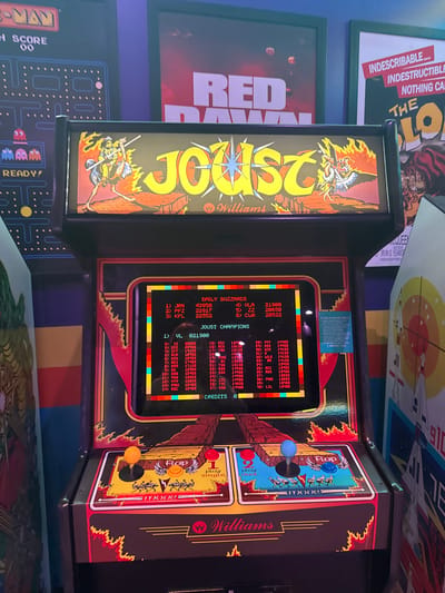 The image showcases a vintage "Joust" arcade machine with vibrant artwork on its sides. The screen displays high scores from "Daily Buzzards" and "Joust Champions," listing player initials and scores. The control panel features two sets of joysticks and buttons for two-player action. The surrounding area includes framed posters of "Red Dawn," a Pac-Man machine, and another partially visible poster, creating a retro arcade ambiance.