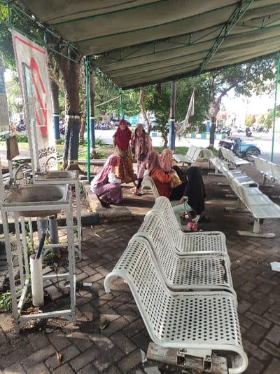 This outdoor image captures a public waiting area in Situbondo, Indonesia, under bright daylight on what appears to be a clear, sunny day. A green canvas canopy provides shade over a brick-paved ground.

The scene features several women, most of whom are wearing hijabs in various colors, including light brown, maroon, reddish-purple, black, and light purple/pink. They are seated on or near white, perforated metal benches, appearing to be resting or waiting. One woman in a light green top is partially visible behind others.

In the foreground, a utilitarian hand-washing station is prominent. It consists of a metal frame supporting a basin and is equipped with PVC pipes, suggesting a foot-operated water dispenser. Adjacent to this station, a white sign displays a red, stylized logo with faint, unreadable text below it. Another small, unreadable sign is visible nearby.

The white perforated metal benches, characteristic of public facilities, are arranged in rows, with some occupied and others empty. The background reveals trees and greenery, hinting at a natural environment beyond the immediate waiting zone. Distant motorbikes and a blue and white striped barrier suggest the presence of a nearby road or parking area. The overall atmosphere is calm and passive, typical of a public gathering or waiting point.