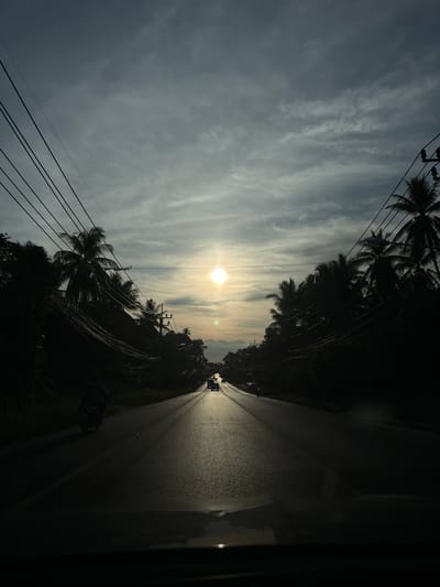 The image depicts a road in Baan Lipa Noi, Thailand, at sunset. The sun, a bright orb, is positioned high in the sky, casting a warm glow over the scene. Palm trees line both sides of the road, their silhouettes creating a dramatic contrast against the colorful sky. A few cars and a motorcycle are visible on the road, suggesting normal traffic activity. Overhead, a network of power lines stretches across the frame, adding a touch of urban infrastructure to the tropical landscape. The overall impression is one of a peaceful, scenic drive through a tropical paradise.