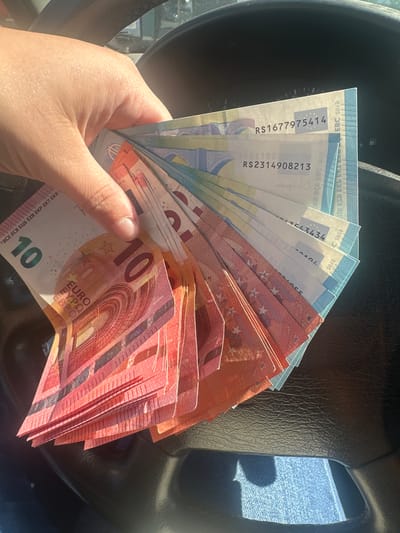 A hand holds a fan of Euro banknotes, comprised of ten-Euro, twenty-Euro, and five-Euro denominations, against the backdrop of what appears to be the interior of a vehicle, with a steering wheel visible. The denominations are mixed and the arrangement suggests a collection of various values of European currency. There is no explicit information to place this image in the city of Rakovski, Bulgaria.