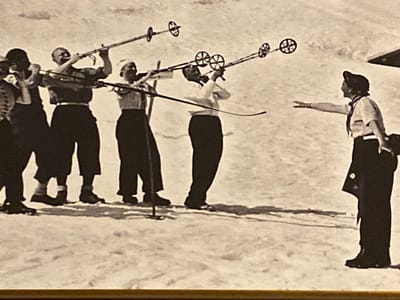 In a snow-covered outdoor environment, a group of six individuals is depicted. Five people are positioned in the left and center of the frame, all wearing ski attire and holding ski poles. The leftmost individual is partially obscured. The four individuals in the middle are facing towards the right, with ski poles held aloft in front of them. Their stances suggest they might be engaged in a coordinated activity or demonstration. To the right of this group, a sixth person, also in ski attire and wearing a hat, is facing left and extending their right arm towards the group. This person's posture and extended arm imply an interaction or instruction directed at the other individuals. The environment appears to be a snowy landscape, possibly a ski slope, with subtle shadows cast on the snow, indicating a bright light source. The image is monochromatic, characteristic of older photography. The presence of skis and poles strongly suggests a winter recreational or training setting.