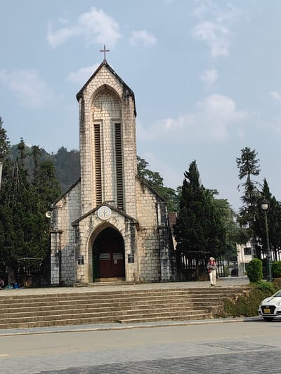 The image captures the Sa Pa Stone Church (Holy Rosary Church) in Sa Pa, Vietnam, on a clear day with scattered clouds. The church, constructed from light-colored stone blocks, features a central bell tower with arched belfry openings and an ornate cross atop its pointed roof. A circular emblem is positioned above the main arched entrance, which contains dark wooden doors and is flanked by informational plaques. A white horn speaker is mounted on the left side of the church's facade.
A wide flight of stone steps leads up to the church entrance from the foreground. Below the steps, a paved road spans the lower portion of the frame, with a tiled sidewalk section on the left. On the far left, at the base of the church steps and beneath a tall evergreen tree adorned with string lights, two individuals are seated on the ground; one has a blue bag nearby. On the right side, a person wearing light-colored pants, a grey top, and a light head covering stands on the upper steps, facing left and carrying a shoulder bag.
Numerous evergreen trees, some decorated with string lights, densely surround the church. A tall streetlamp with an opaque globe is visible on the right. In the distant background, forested hills rise. A white Hyundai car with a Vietnamese license plate (24A 105.85) is partially visible on the right side of the road.