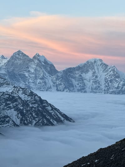 This image captures a majestic high-altitude mountain landscape, likely at either sunrise or sunset, above Khumjung, Nepal. The scene is dominated by rugged, snow-capped mountains emerging from a thick blanket of clouds, creating a dramatic cloud inversion.
The sky, occupying the upper part of the frame, displays a soft gradient of colors. It transitions from a light, clear blue at the very top to warm streaks of pink, orange, and pale lavender, indicating the atmospheric conditions typical of dawn or dusk.
Below the colorful sky, imposing mountain peaks with dark, rocky faces and patches of snow and ice pierce through the cloud layer. A prominent, dark, craggy peak stands on the right side, its steep slopes contrasting sharply with the bright clouds. Further into the distance, other lighter blue-grey mountains with visible glaciers and snowfields are partially visible, rising from the extensive cloud bank.
A dense, seemingly endless "sea of clouds" or fog covers the lower and middle ground, creating a vast, flat, and ethereal surface from which the mountain summits emerge. The top surface of this cloud layer appears bright and diffused, with subtle textural variations. The overall atmosphere is serene and awe-inspiring, highlighting the grandeur of the Himalayan environment. There is no visible human activity or text in the image.