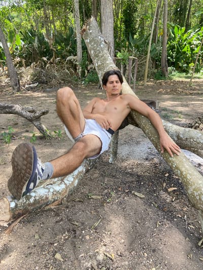 A shirtless man with dark hair and a light complexion is reclining on fallen tree branches in a wooded area. He is wearing white shorts and grey socks with blue and black athletic shoes. His left leg is extended, and his right arm is resting along a branch. The ground is covered in dirt, small stones, and fallen leaves. In the background, there are trees and dense vegetation. The lighting suggests it is daytime with sunlight filtering through the trees. The scene is outdoors in a natural environment, possibly a park or forest area within Los Velásquez, Venezuela.