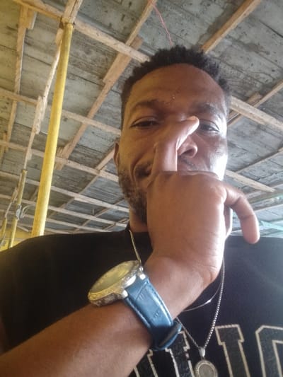 The image is a close-up selfie of a man, taken from a low angle, looking upwards into the camera. The man has dark skin, short dark textured hair, and a neatly trimmed beard and mustache. His right hand is raised, with his index finger resting on the bridge of his nose and his thumb positioned near his mouth, partially obscuring the lower half of his face. He appears to be smiling or smirking, with his left eye visible and showing a hint of amusement.

He is wearing a dark, possibly black, t-shirt. On his left wrist, he sports a prominent watch with a large, circular, silver-toned casing. The watch bezel appears to be encrusted with small, reflective stones, and it has a dark blue, textured strap, likely made of leather or faux leather. Around his neck, he wears a silver-toned chain with a circular pendant that features an intricate, possibly embossed or engraved, design in its center.

The background shows an overhead structure made of wooden beams and planks, forming a ceiling or roof. Some yellow-colored poles or supports are visible on the left side of the frame, suggesting a rustic or semi-finished indoor or covered outdoor setting, possibly a shed, construction area, or an open-air sheltered space. The lighting indicates it is daytime. No discernible text is visible in the image.