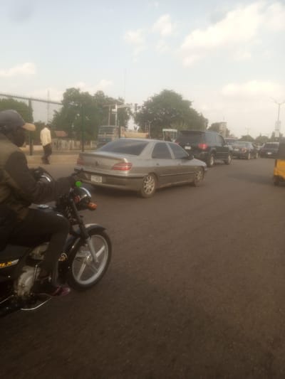 A street scene in Unguwan Dosa, Nigeria, features a motorcycle rider in the foreground, wearing a dark cap and jacket, with a glimpse of their striped footwear. The rider is positioned on a black motorcycle with a silver wheel rim. Behind the motorcycle, traffic is visible on a paved road. A silver sedan is prominent in the middle ground, displaying a license plate that appears to read "CRC 866A". Further back, other vehicles, including a dark SUV and more sedans, are part of the traffic flow. In the background, trees and buildings suggest an urban or semi-urban environment. The sky is cloudy, indicating it might be late afternoon or early morning. The lighting suggests natural daylight, possibly during a hazy or overcast period. The overall impression is a snapshot of daily life and transportation in a Nigerian town.