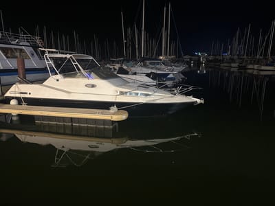 At night, several boats are docked in a marina in Agde, France. The foreground features a white motorboat with a black hull, its reflection visible in the dark water. Behind it, numerous sailboats with tall masts are moored. The marina is illuminated by artificial lights, creating reflections on the water's surface. In the background, distant lights suggest the presence of buildings or structures on land. The overall scene depicts a tranquil night at a French marina.