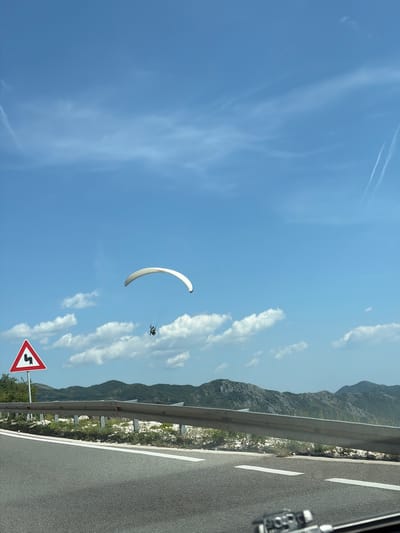 The image captures a paraglider suspended in a bright blue sky over the scenic landscape of Brajici, Montenegro. The paraglider, a person in a yellow and black suit, is positioned under a large white canopy, appearing to be mid-flight. Below the paraglider, a winding road is visible, bordered by a guardrail and sparse vegetation. A triangular road sign with a zigzag symbol indicating a winding road is present on the left side of the frame, near a cluster of green trees. In the background, a range of rugged, green mountains stretches across the horizon, partially obscured by a few scattered white clouds. The overall scene suggests an outdoor recreational activity taking place on a clear, sunny day in a mountainous coastal region.