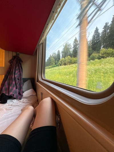 The image shows the interior of a train carriage, looking out the window at a rural landscape. The window is large and curved, with a tan-colored frame. Through the window, a field of white wildflowers is visible, with a line of trees in the background. A utility pole with wires runs through the scene outside. Inside the carriage, a red ceiling is visible above the window. A plaid shirt is hanging on a hook to the left of the window. In the foreground, a person's legs are visible, wearing black shorts. The bed or seat of the carriage is covered in white sheets. The lighting suggests it is daytime. The scene is consistent with travel through a rural area, potentially near Pokrovka, Russia, given the mention of that location.