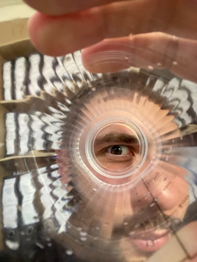 The image is a first-person perspective, looking through the opening of a plastic container. A person's right eye is visible, centered in the opening, with a brown iris and dark pupil. The person's mouth and nose are also partially visible, with pink lips. The container has circular ridges and some markings that appear to be measurements. In the background, out of focus, are shelves containing objects with black and white stripes, possibly books or boxes. The lighting suggests an indoor environment. The location is Luzhitsy, Russia.