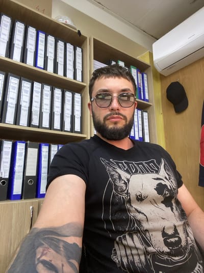 In the city of Luzhitsy, Russia, a man with a beard and sunglasses sits in an office, facing the camera. He is wearing a black t-shirt with a graphic print of a dog's head. To his left, a wooden bookshelf is filled with binders. Above him, an air conditioning unit is mounted on the wall. To his right, a black baseball cap hangs on the wall. His left arm, visible in the foreground, is covered in a large tattoo. The overall environment appears to be a typical office setting with ample storage.