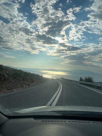 The image captures a view from inside a vehicle driving on a coastal road in the Nea Makri Municipal Unit, Greece. The road curves to the right, with a steep, rocky embankment on the left covered in sparse vegetation. On the right, a metal guardrail separates the road from a vast expanse of water, likely the Aegean Sea, which reflects the bright sunlight filtering through the clouds. Distant landmasses and mountains are visible on the horizon. The sky is filled with dramatic, fluffy white and gray clouds, with patches of blue sky peeking through. The sun is partially obscured by clouds, creating a bright, luminous effect on the water. The dashboard of the car is visible in the foreground. There are no people or animals depicted in the scene.
