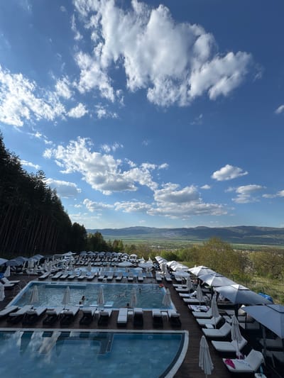 A long rectangular swimming pool surrounded by lounge chairs and white umbrellas is set against a backdrop of mountains and a partly cloudy blue sky. The location is likely a resort or hotel in Sapareva Banya, Bulgaria. The pool has people swimming in it. Trees border one side of the scene.