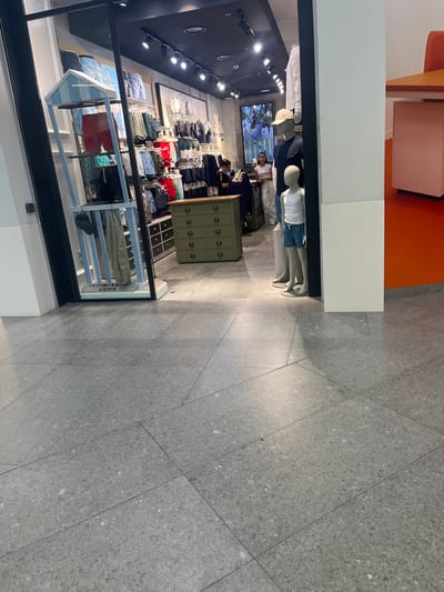 The image shows the interior of a clothing store, likely a men's boutique, situated in Limbiate, Italy. The store features display racks filled with men's shorts and clothing, with a focus on summer wear. A prominent display includes a small, beach-hut-like structure with shorts displayed on shelves. Mannequins are present, one of which is a child-sized mannequin wearing a white tank top and blue patterned shorts. A woman is visible behind a counter in the background. The floor is tiled with large, grey, speckled tiles. The store appears clean and well-organized, with modern lighting fixtures on the ceiling. To the right, a part of a desk or counter with an orange surface is visible, suggesting a checkout or customer service area. The overall impression is of a retail environment in a commercial area.