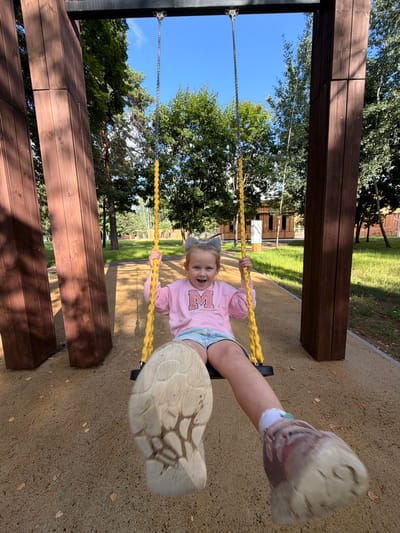 In Malakhovka, Russia, a young girl with grey cat ears on her head is joyfully swinging on a yellow rope swing. She wears a pink sweatshirt with a large 'M' on it and denim shorts. Her right foot is prominently in the foreground, mid-air with a worn sneaker. The swing set has two thick, dark brown wooden posts. The background features a bright blue sky, lush green trees, and a modern-looking wooden building. The ground is covered with sand, with scattered autumn leaves.