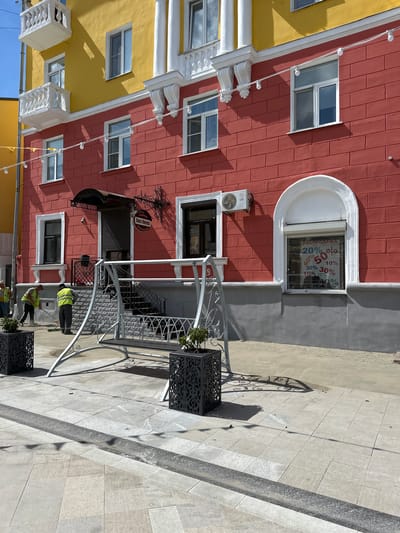 In Putyatino (Semivragovskaya S/A), Russia, a commercial building with a vibrant red facade is depicted under a clear sky. The building features yellow accents on its upper sections and white decorative elements around the windows and balconies. Two men in high-visibility vests are engaged in cleaning activities on the paved sidewalk in front of the building. One man is using a broom, and the other appears to be sweeping. A decorative metal swing is positioned on the sidewalk, and two large, ornate planters are placed on either side of it. A window on the red facade displays a "SALE" sign with various discount percentages. An air conditioning unit is mounted on the wall near one of the windows. The surrounding environment is a sunlit urban setting with paved walkways and shadows cast by the building and the swing.
