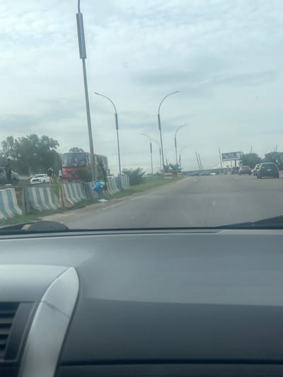 The image shows a view from inside a vehicle looking out at a highway scene in Ankuru, Nigeria. A red bus is parked on the side of the road, and several people are standing near it. A white pickup truck and other vehicles are also visible. The road is lined with concrete barriers with blue and white stripes. Streetlights are spaced along the side of the road. The sky is overcast.