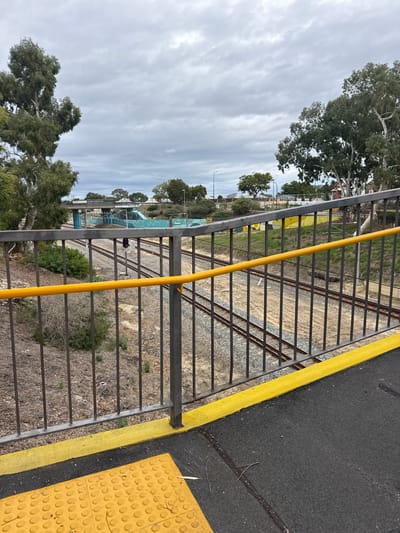 The image depicts a view from a pedestrian bridge overlooking railway tracks in Bassendean, Australia. The foreground features a metal safety railing with a yellow vertical pole. To the right, there is a section of black asphalt with a yellow safety stripe and tactile paving. Beyond the railing, railway tracks are visible, surrounded by gravel and sparse vegetation. In the distance, there is a building with a blue, dome-like structure. Trees are present in the scene, and the sky is overcast. The photograph appears to have been taken during the daytime.