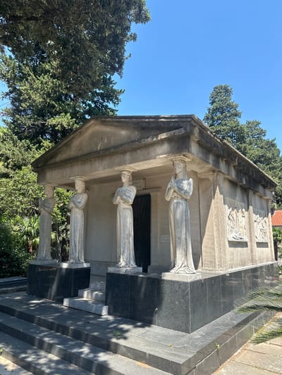 The image shows a tomb in the city of Škaljari, Montenegro. The tomb is a rectangular structure with a triangular pediment and a flat roof. It is made of stone and is adorned with sculptures of women. The sculptures are in a classical style and are depicted in draped robes. The tomb is surrounded by trees and foliage. The sky is clear and blue. There are no people in the image. The tomb is located on a hill, and there are steps leading up to it. The overall scene is one of solemnity and grandeur.