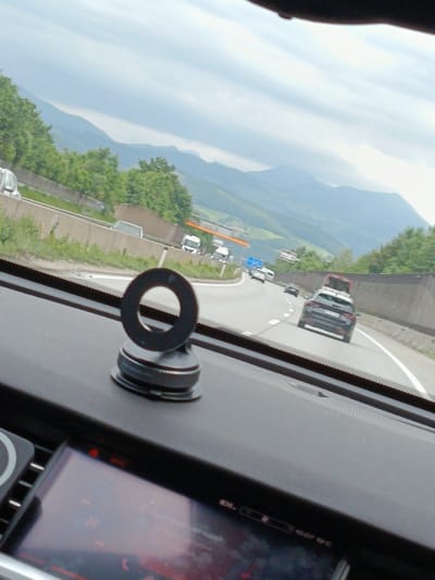 The image is a first-person perspective from inside a vehicle on a highway, likely traveling through a mountainous region. In the foreground, on the dashboard, is a circular object with a base, possibly a phone mount. The dashboard itself is dark. The windshield shows a highway with several cars and trucks. The landscape outside features green trees and rolling hills leading to distant mountains under a cloudy sky. The specific location in Glanegg, Austria, cannot be determined from the visual information presented. There are no people clearly visible. The overall scene depicts transit on a highway.