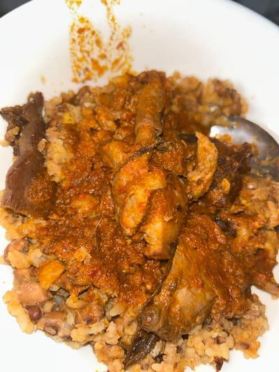 A white bowl contains a serving of food. The base layer consists of a granular mixture, appearing to be rice and beans, with a light brown and beige coloration, interspersed with darker bean specks. This base is covered by a thick, reddish-orange stew, rich in texture and color, indicative of a tomato and chili pepper base. Embedded within the stew are various pieces of protein, including irregular chunks of lighter-colored meat, possibly poultry or red meat, and darker, elongated pieces that may be dried fish or offal. A silver-colored spoon is partially submerged in the food on the right side of the bowl. Splatters of the reddish-orange stew are visible on the upper interior rim of the white bowl. No people or other environmental details are visible.