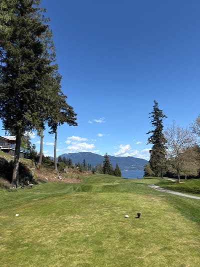 The image depicts a golf course in Bowen Island Municipality, Canada, on a clear day with a bright blue sky and scattered cumulus clouds. The green of the golf course occupies the right side of the frame, leading towards mountains and a body of water in the background. On the left, tall evergreen trees frame the view, with a partial view of a house at the bottom. A golf ball sits on a tee near a black tee marker on the green. In the distance, snow-capped mountains are visible.
