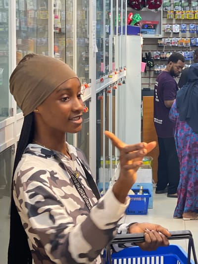 A young Black woman wearing a brown headscarf and a camouflage hoodie is standing in a shop in Guzape, Nigeria. She is looking to her right and gesturing with her right hand, which is illuminated by sunlight. She is also holding the handle of a blue shopping basket, which contains various items. Behind her, shelves are filled with merchandise, and other shoppers are visible in the background. The shop appears to be a general store or market.