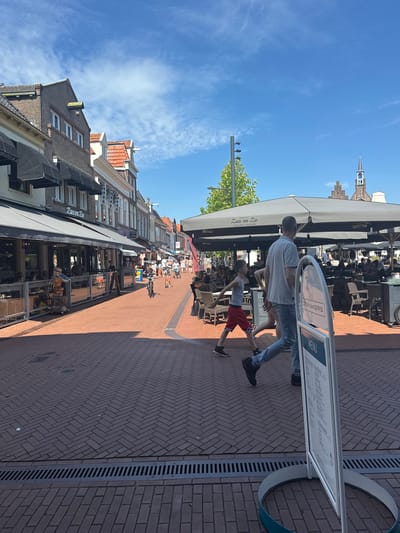 This image depicts a street scene in Steenwijk, Netherlands, likely a town center with shops and restaurants. The foreground is dominated by a red brick paved street laid in a herringbone pattern, with a metal drain grate running horizontally across the lower portion of the frame. A white A-frame sign with a menu is prominently positioned on the right, displaying the word "MENU" and illegible text.
In the middle ground, a man and a young boy are walking from right to left. The man is wearing a light blue t-shirt and jeans, and the boy is wearing a grey tank top and red shorts. They are walking past an outdoor seating area of a restaurant, which features large umbrellas and tables with chairs. Further down the street, other pedestrians are visible, some walking and others appearing to be dining or shopping. Buildings line both sides of the street, with awnings extending over storefronts. One awning has the text "Zus en Zo" visible. A church steeple with a clock can be seen in the distance on the right. The sky is blue with scattered white clouds. The overall impression is a bustling urban environment on a sunny day.