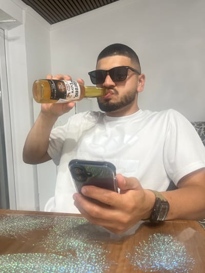 A man is shown from the chest up, seated at a table with a holographic glitter pattern. He is wearing a white t-shirt, sunglasses, and a watch. He is holding a bottle of Corona beer in his left hand, drinking from it, and simultaneously holding a smartphone in his right hand. The background consists of a white wall and a section of a white ceiling with wooden slats. The image appears to be taken indoors, possibly in a residential setting in Rosenovo, Bulgaria.