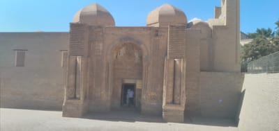 The image shows the Chor Minor Madrasah in Bukhara, Uzbekistan. The madrasah is made of brick and has a distinctive dome. The entrance to the madrasah is a large archway with intricate carvings. A person in a white shirt is seen entering the madrasah. The sky is clear and blue.