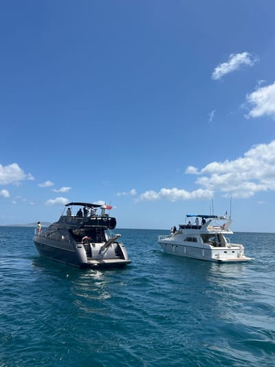 The image depicts two yachts on the water under a blue sky with scattered white clouds. The yachts are positioned side by side. One is a predominantly white vessel, while the other is dark gray or black. Both boats appear to have people on board. The yachts are situated near the city of M'Diq, Morocco. The water is a shade of blue-green, and the overall composition is bright and sunlit.