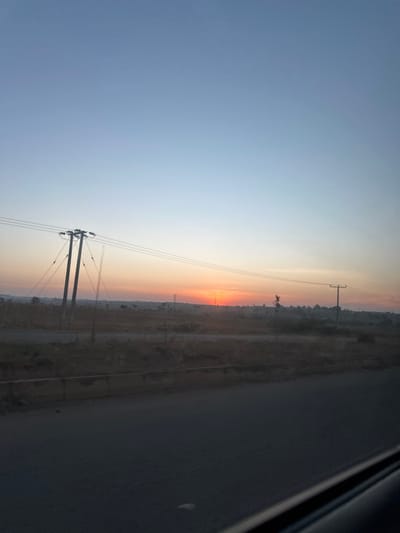 A landscape under a soft sunset sky. Power lines stretch across the horizon from a pole in the foreground. The right side shows a glimpse of a road. The horizon features a muted gradient of oranges and yellows, blending into the light blue sky. Sparse vegetation and shrubs are visible along the horizon line. The overall atmosphere is tranquil.