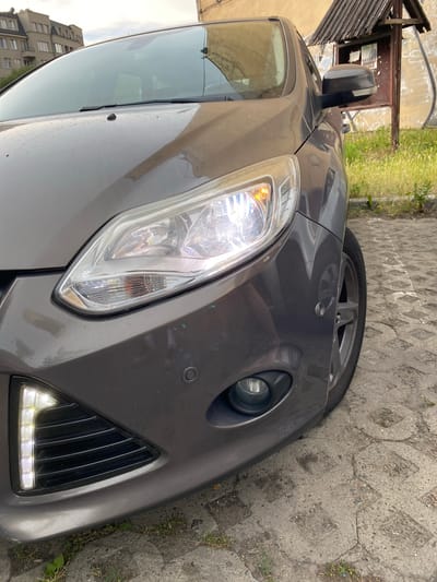 This image shows the front passenger side of a dark gray car, likely a hatchback, parked on a textured gray pavement. The car's headlight is illuminated, revealing a clear lens and bright white and orange lights. To the left of the headlight, a black grille with vertical slats is visible, with a strip of LED daytime running lights above it, also illuminated. A circular parking sensor is embedded in the bumper below the grille. A fog light is also visible on the lower part of the bumper.
The car is parked outdoors, with a grassy area and a wooden structure visible to the right. In the background, a multi-story building with visible windows suggests an urban or suburban setting. The lighting suggests it is daytime, with overcast or cloudy skies. The image is taken from a low angle, focusing on the car's front end. There is no visible text in the image. The scene appears to be a still moment, with no activity taking place.