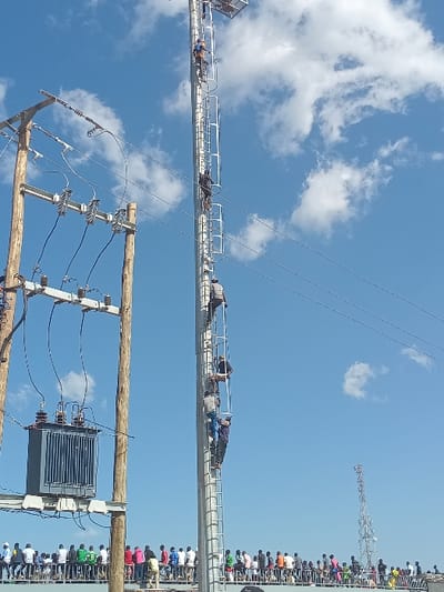 This media file captures an outdoor scene in Siaya, Kenya, under a bright blue sky dotted with scattered white clouds. The image is oriented vertically, making the horizon appear on the left and right sides of the frame.
In the mid-ground, a long, elevated metal structure, resembling a large beam or truss with a railing system, extends horizontally across the view. Along this structure, several individuals are visible; some are lying prone, seemingly resting or watching, while others are sitting, using it as an improvised viewing platform.
To the far right, a large gathering of people is seated in what appears to be bleachers or spectator stands. They are facing away from the camera, dressed in diverse casual attire, suggesting their attendance at an event. In the distant background, a tall metal lattice tower or antenna structure is visible.
In the foreground, on the lower left, a prominent wooden utility pole supports multiple black power lines and a grey electrical transformer. These power lines crisscross portions of the sky.
The scene depicts a daytime event or gathering, with attendees utilizing both conventional seating and an elevated structural element for observation. The weather is clear and sunny. No visible text is present in the image.
