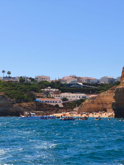 The image depicts a vibrant coastal scene in Lagoa, Portugal, on a clear, sunny day. The bright blue sky and sparkling turquoise water suggest fair weather. The foreground is dominated by the dynamic, rippling surface of the ocean, with whitecaps indicating a gentle breeze.

In the middle ground, a tour boat with the visible text "MARA" emblazoned on its side is filled with people, likely tourists enjoying a boat excursion. The boat is cruising on the water, close to the shore. Several smaller vessels, including kayaks, are also scattered in the water, with people paddling and enjoying various water activities.

The background reveals a picturesque cove with a sandy beach crowded with sunbathers under colorful umbrellas. The beach is backed by dramatic golden cliffs and a hillside dotted with whitewashed buildings with red-tiled roofs, characteristic of Mediterranean architecture. Lush green vegetation clings to the slopes, contrasting with the rocky cliffs. A few palm trees are visible on the higher ground, further adding to the tropical feel. The scene conveys a sense of leisure and vacationing, with people actively engaged in enjoying the beautiful coastal environment. The overall impression is one of a lively tourist destination during peak season.