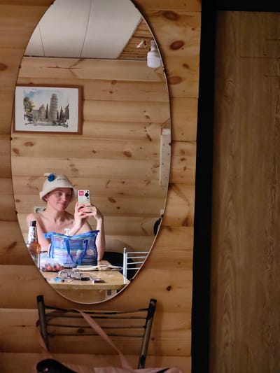 In an indoor scene with wood-paneled walls, a woman in a white sauna hat with a blue flower is reflected in an oval mirror. She is holding a smartphone and taking a selfie, with her left arm bare and her right arm holding the phone. On the wooden table in front of her reflection, there is a bottle of beer, a transparent blue striped bag, a plastic bag filled with cherries, a pair of glasses, and a set of keys. A framed watercolor painting of the Leaning Tower of Pisa hangs on the wall above and to the left of the mirror. A metal chair with a light pink strap is visible in the foreground. The overall environment suggests a rustic or traditional setting, possibly a sauna or a cabin.